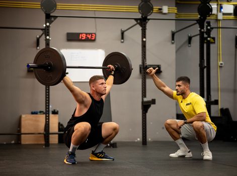 Athlete performing weightlifting exercise under trainer's guidance in a gym setting.