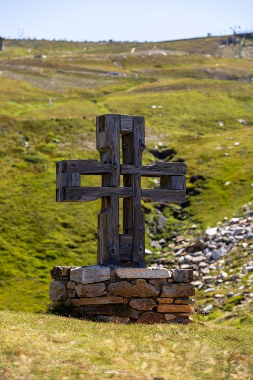 Free A wooden cross stands on a stone base in a grassy hill with vibrant green grass under clear skies. Stock Photo