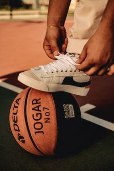 Man tying sneakers on a basketball at an outdoor court, emphasizing sports and activity.