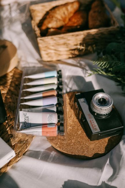 A vintage camera sits beside acrylic paints on a sunny outdoor surface.