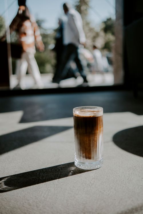 Free A refreshing latte in a glass catches the sunlight in an urban café, with blurred pedestrians in the background. Stock Photo