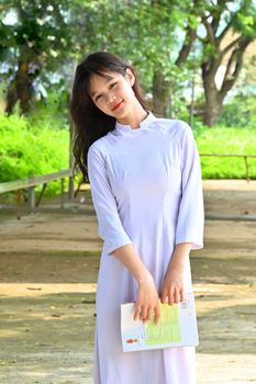 Portrait of an Asian woman in a traditional white Ao Dai, standing outdoors with a book.