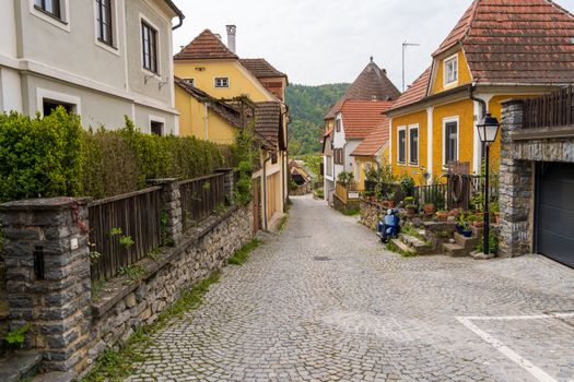 Picturesque narrow street in a European village with cobblestone, colorful houses, and lush greenery.