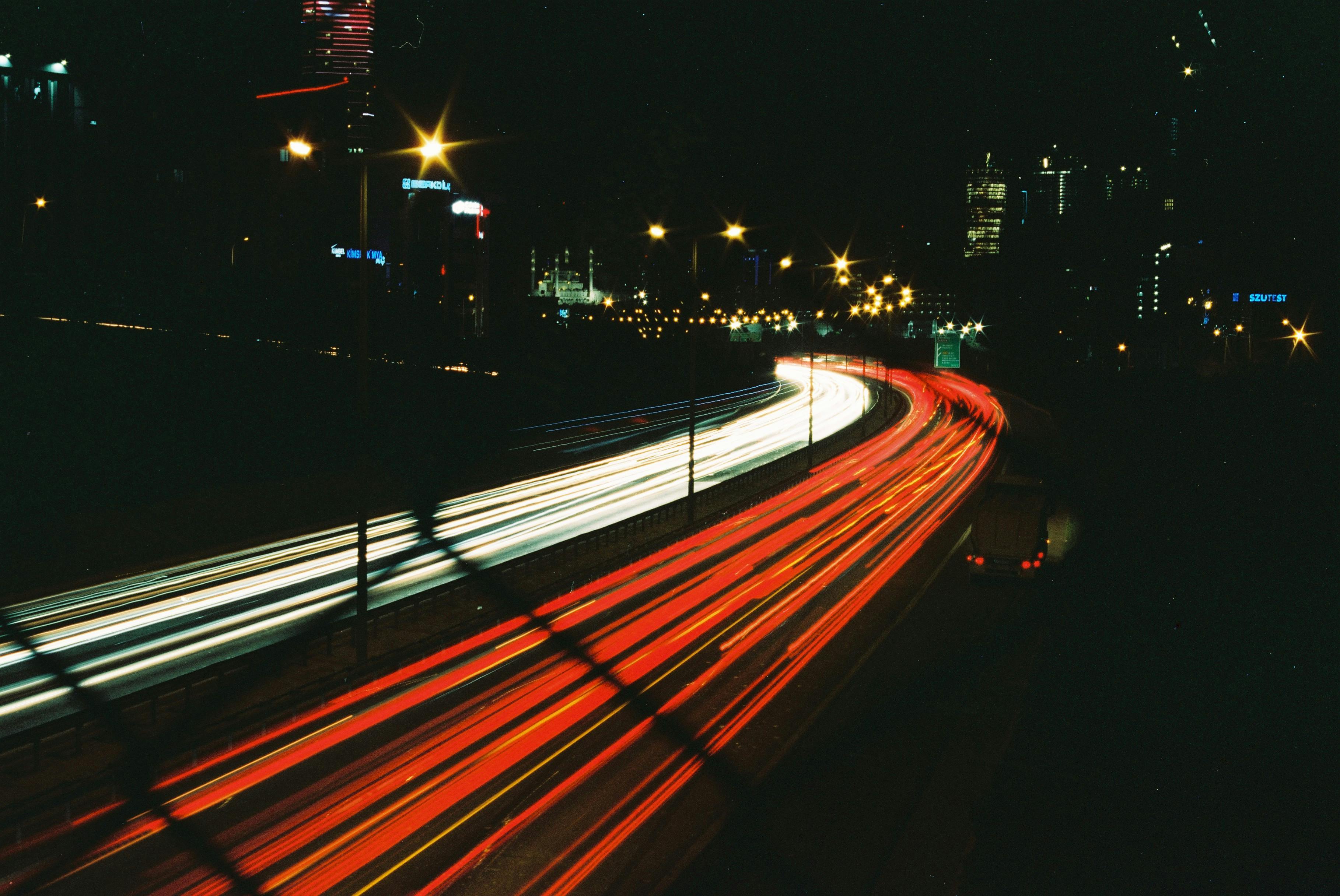 Long exposure of bustling city highway at night with vibrant light trails and illuminated buildings.