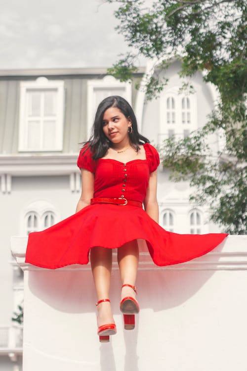 Model in a Red A-Line Dress and High Heels Sitting on a White Wall