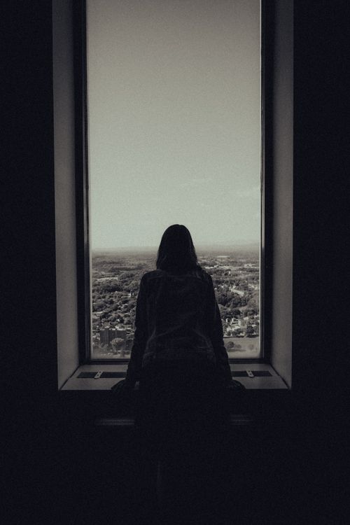 Free Silhouette of a woman standing by a large window overlooking a cityscape, creating a reflective mood. Stock Photo