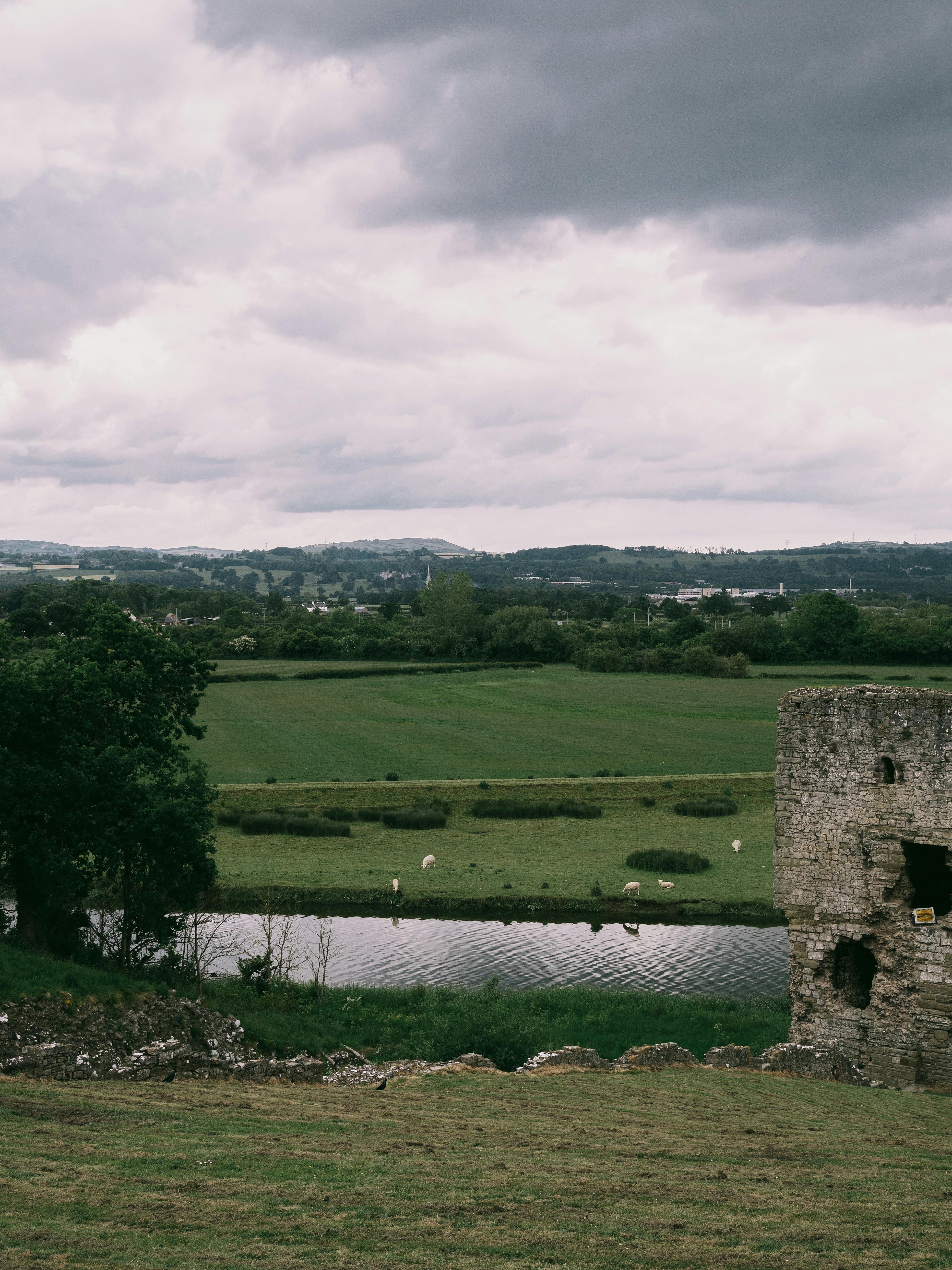 Tranquil countryside view featuring pastures, canal, and ancient stone ruins under an overcast sky.