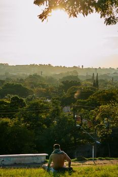Shirtless man sitting outdoors enjoying a sunny day, overlooking a lush green landscape.