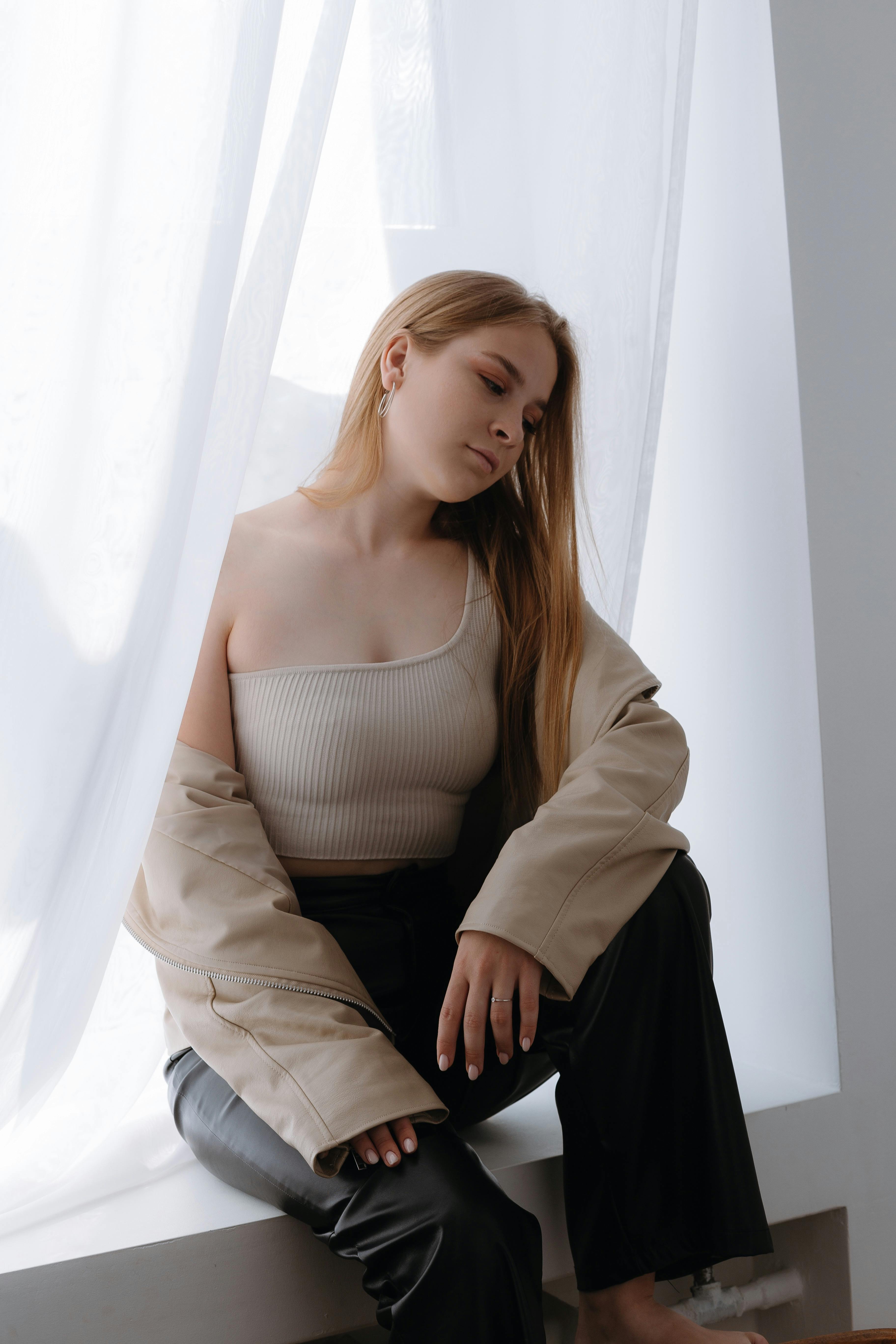 Free Elegant woman in beige outfit sitting by a windowsill. Fashion photography with soft lighting and serene mood. Stock Photo