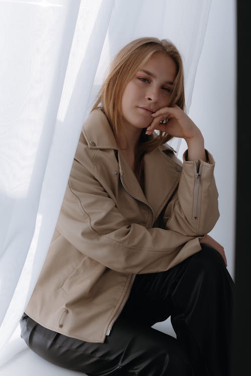 Blonde Woman in Beige Leather Jacket and Black Pants Sitting on Windowsill