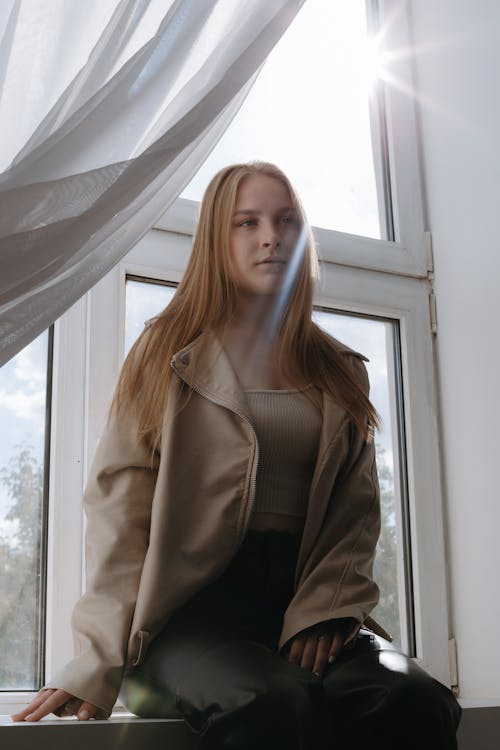 Blonde Woman in Beige Leather Jacket Sitting on Windowsill 