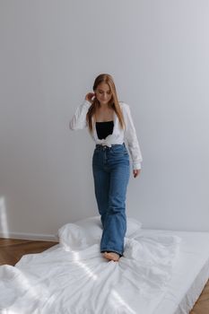 Young woman in casual attire standing on a bed in a minimalistic room.