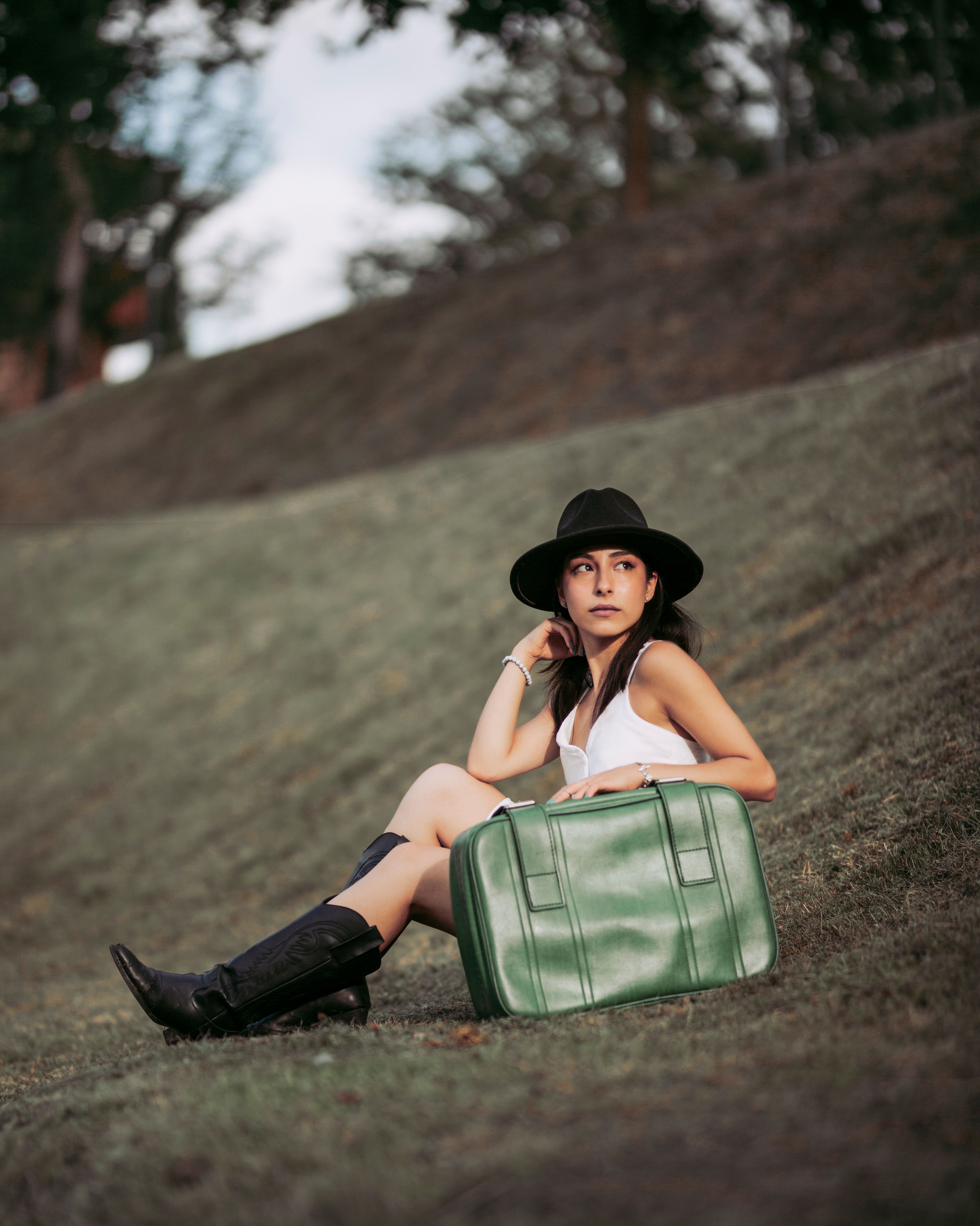 Fashionable woman sitting outdoors with a green suitcase, wearing a hat and boots.