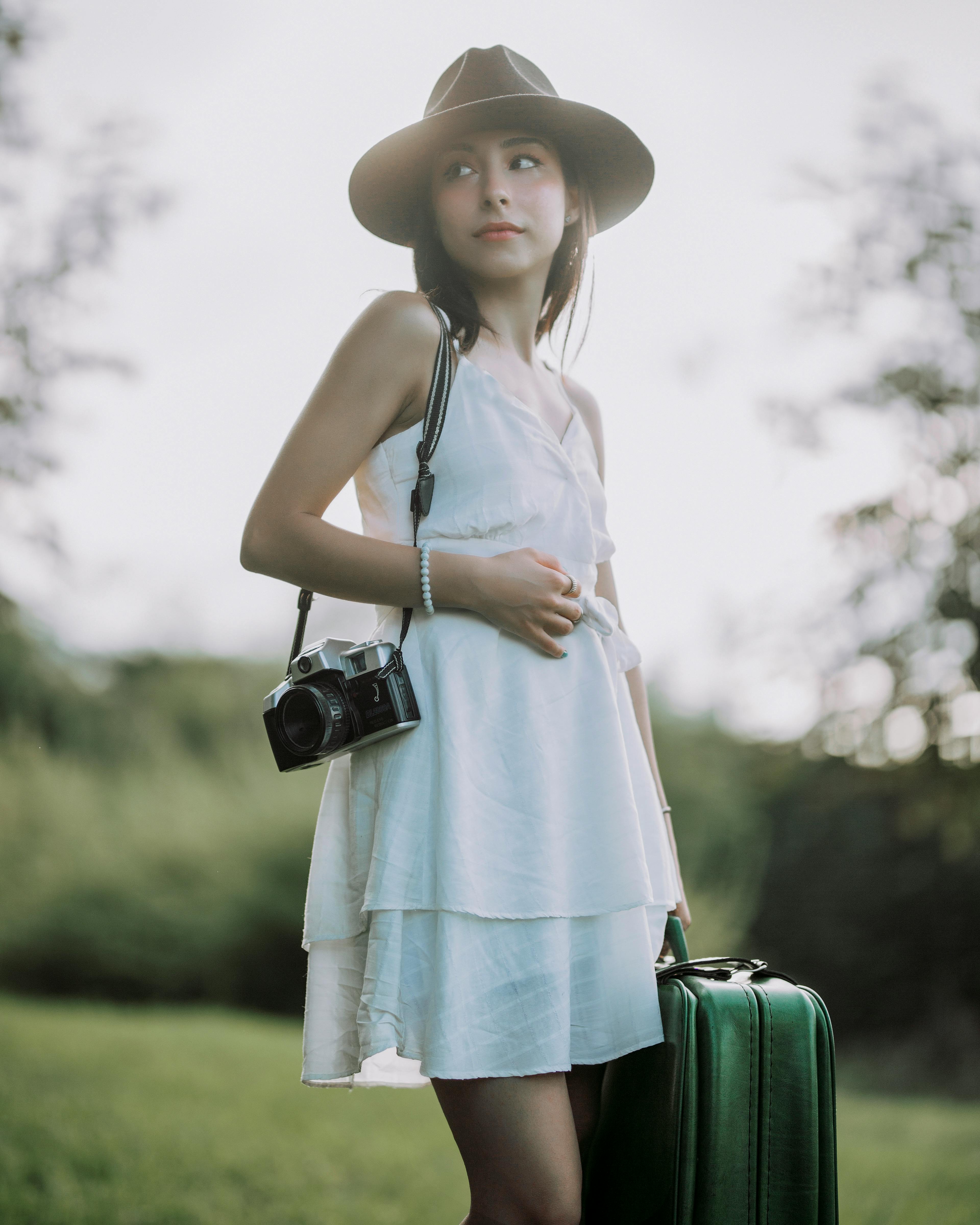 Young woman with hat and vintage camera posing outdoors with suitcase.