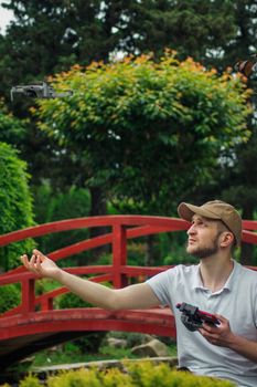 A young man navigates a drone near a red bridge surrounded by vibrant greenery.