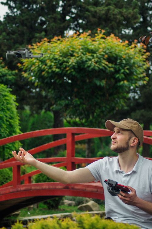 Free A young man navigates a drone near a red bridge surrounded by vibrant greenery. Stock Photo