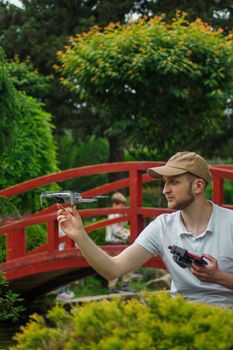 Man flying a drone beside a red bridge in a lush green park.