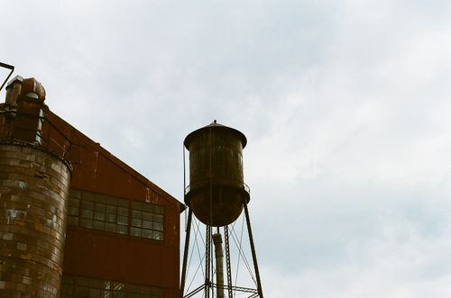 Free A rustic water tower and industrial building under a cloudy sky, showcasing urban decay. Stock Photo