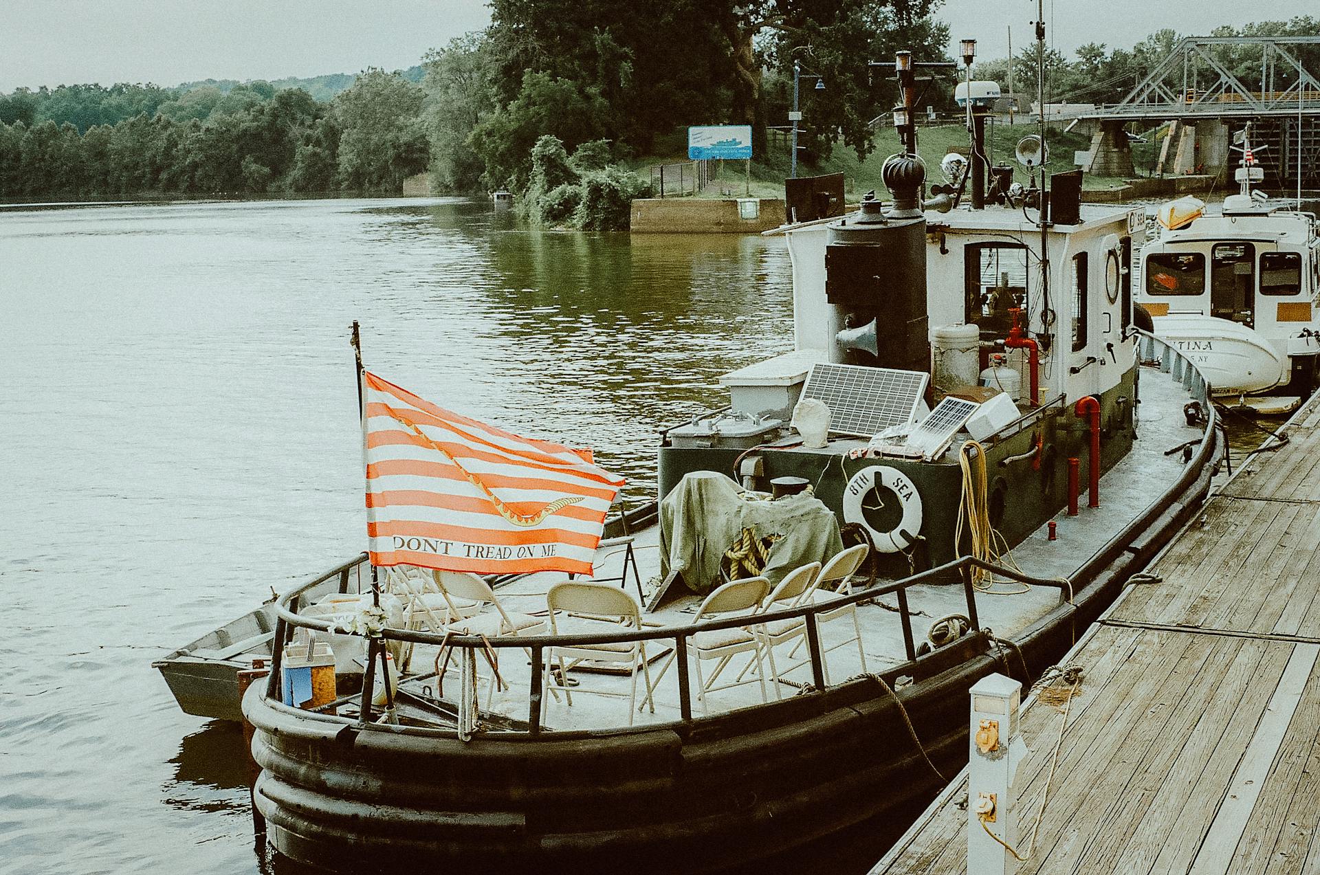 A rustic tugboat with a 'Don't Tread on Me' flag docked on a serene river by a pier.