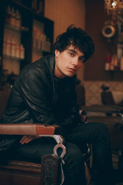 Fashionable young man posing in a stylish black outfit indoors, captured in a moody setting.