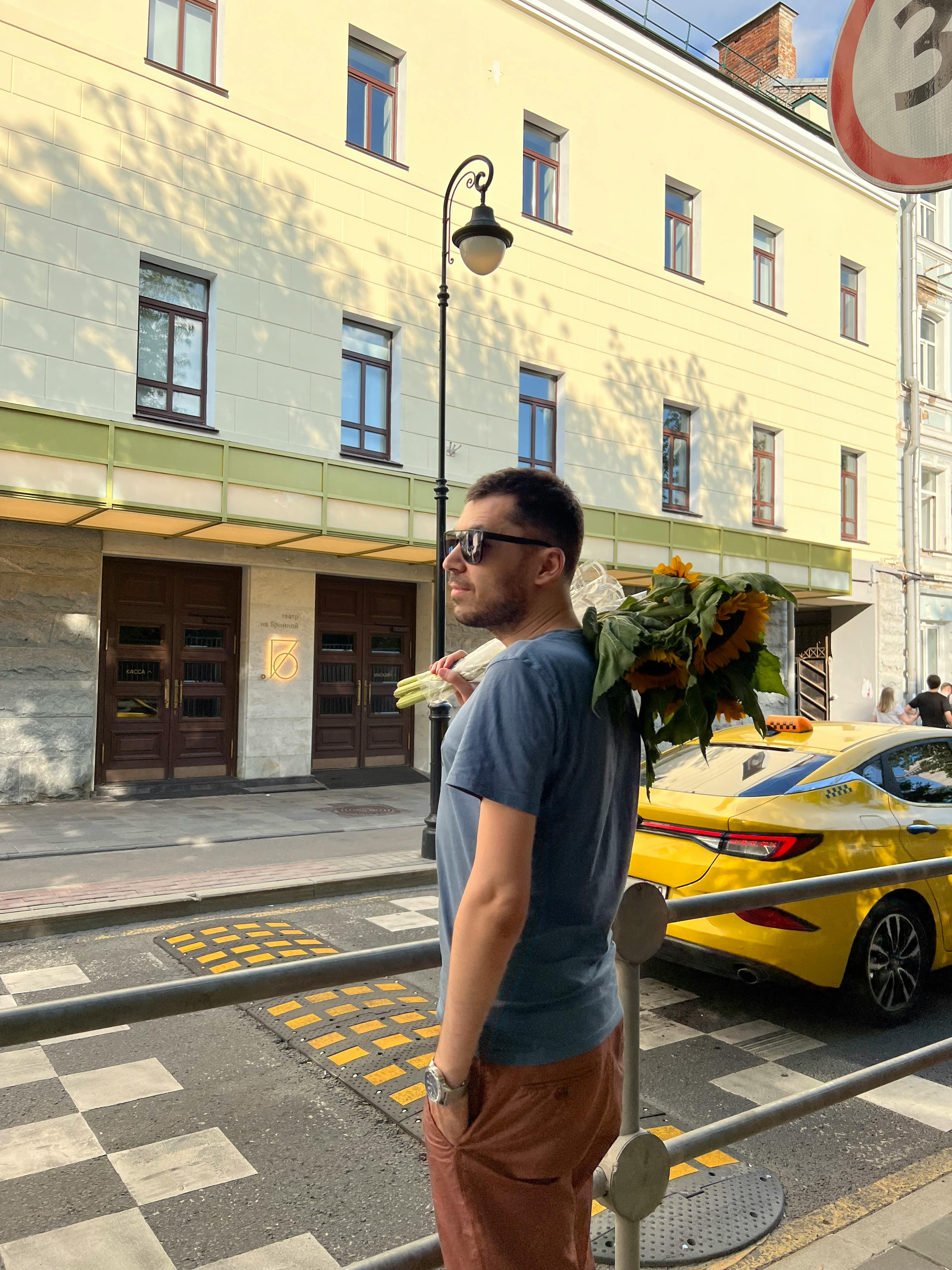 A man stands in an urban street holding a vibrant bouquet, near a yellow taxi on a sunny day.