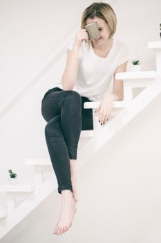 Young joyful barefooted female in casual clothes chilling with legs crossed on white stairs having morning beverage