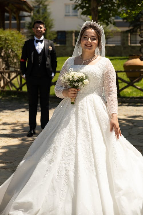 Smiling Woman with Flowers and in Wedding Dress Standing in Park with Man in Suit behind