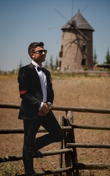 Elegant groom in a tuxedo and sunglasses poses by a rustic fence near a countryside windmill.