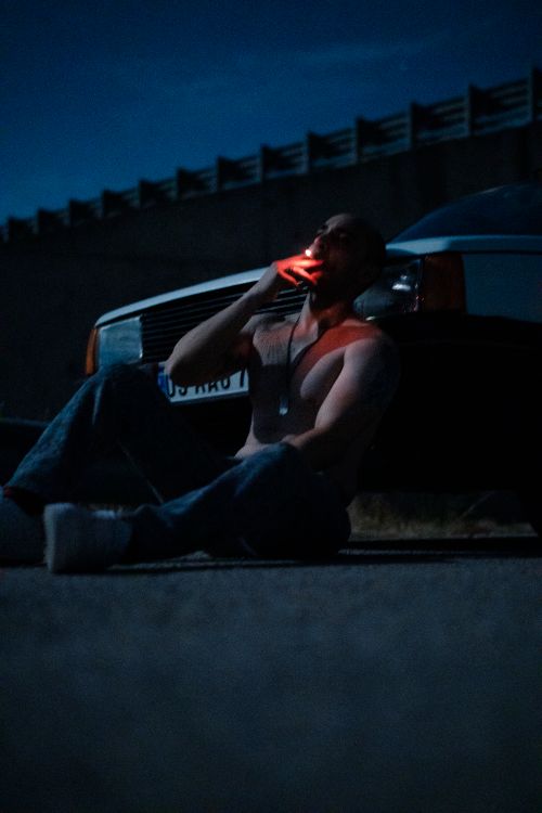 Free A shirtless man sits by a car, smoking at night under a clear sky. Stock Photo