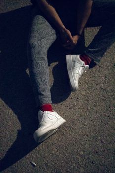 Person sitting on asphalt, showcasing casual fashion with white sneakers and patterned pants in sunlight.