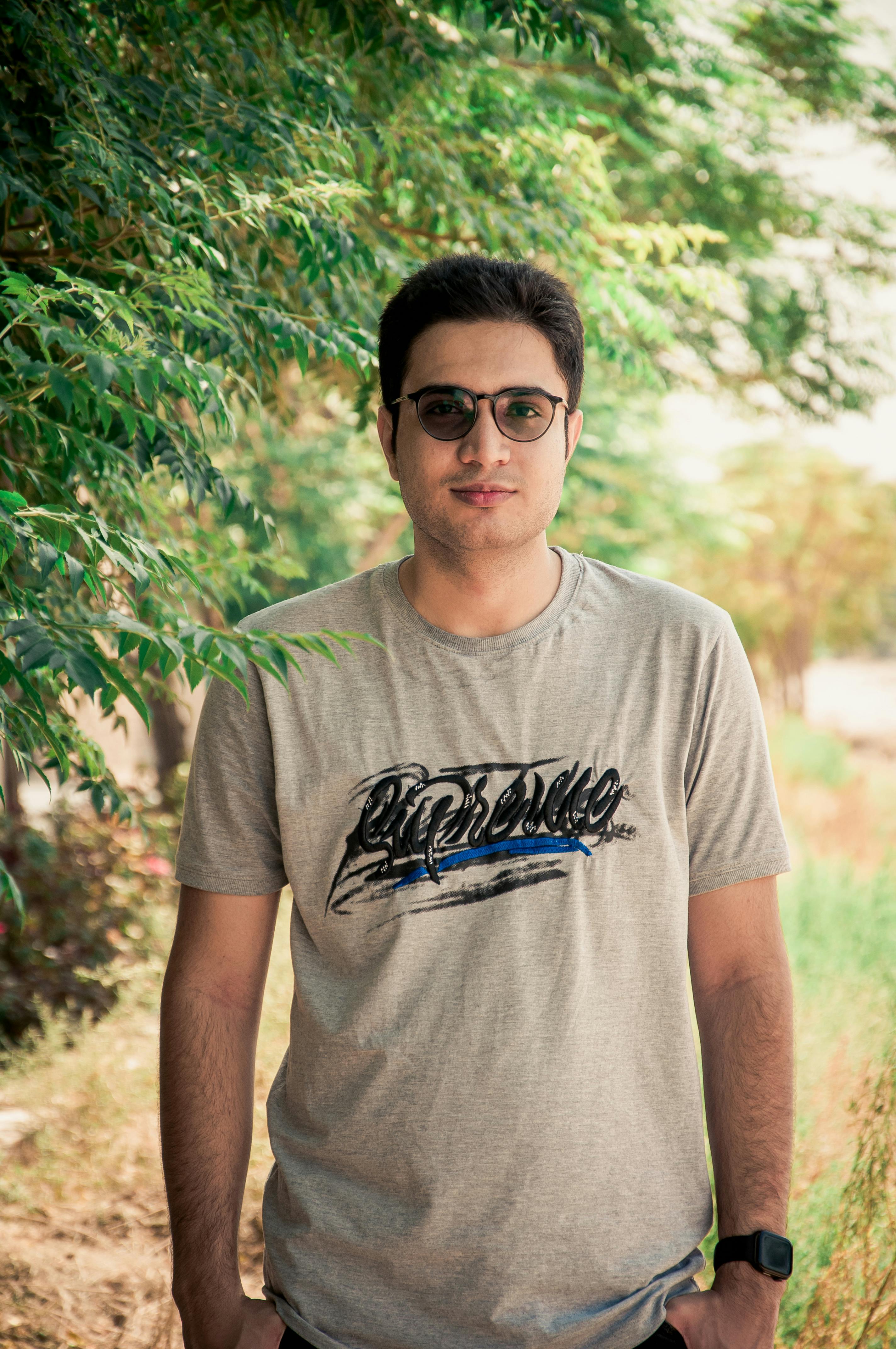 A relaxed portrait of a man standing outdoors in sunglasses, with natural greenery in the background.