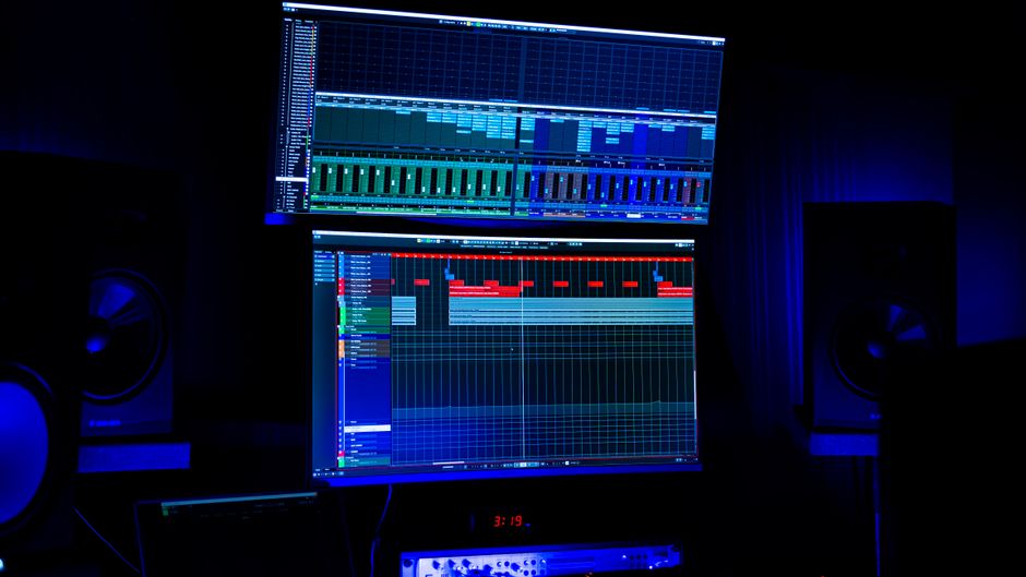 A modern recording studio featuring two screens displaying audio editing software under blue neon lighting.