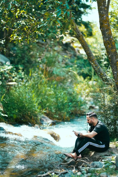 Free A man enjoys nature by a river in a forest while using a smartphone. Stock Photo