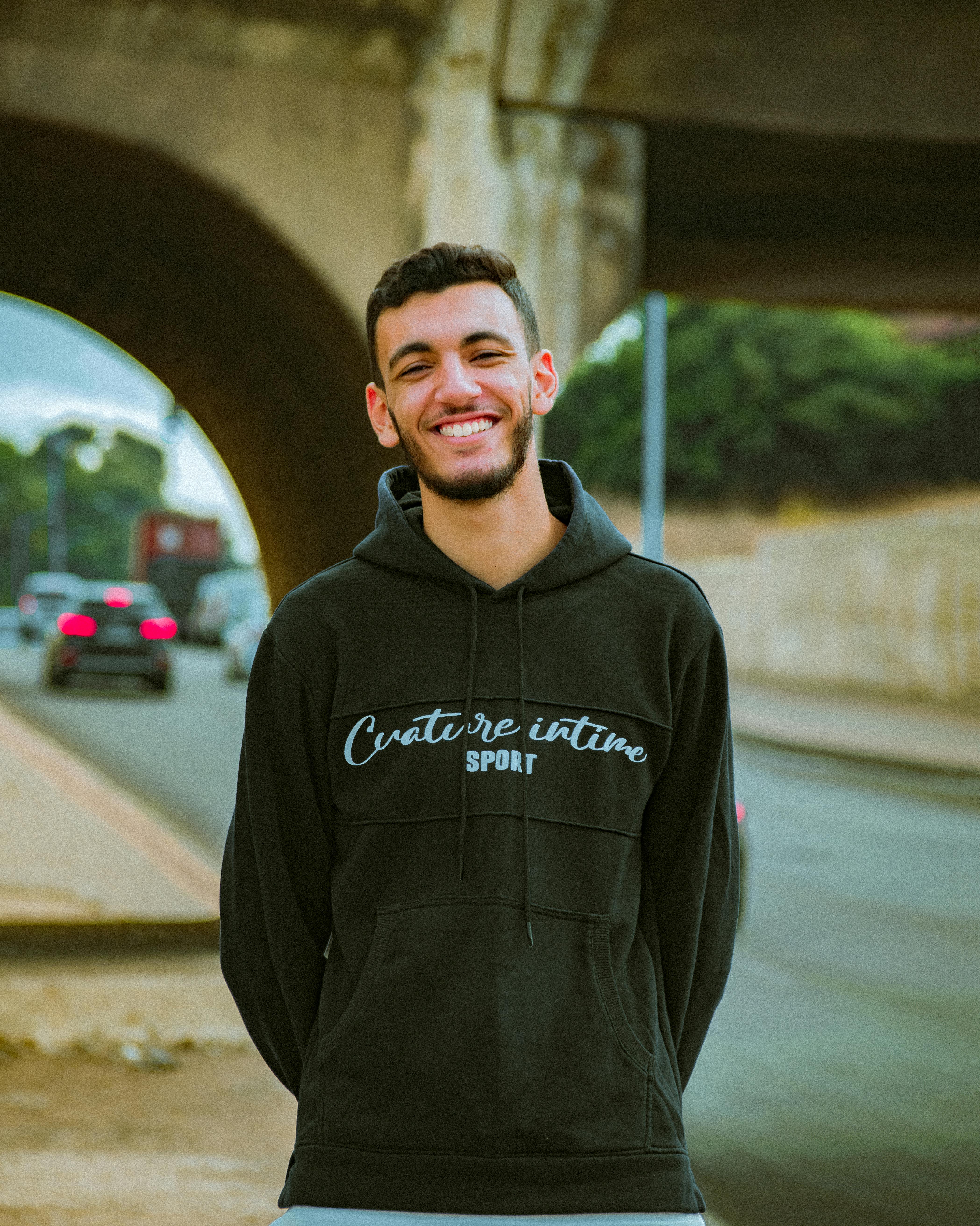Casual portrait of a man in a black hoodie smiling under a bridge in an urban setting.