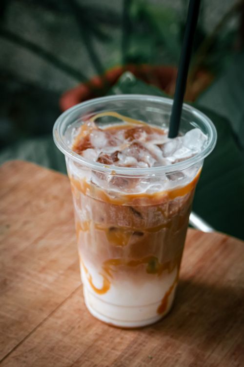 Free Close-up of iced caramel macchiato with straw on wooden table, surrounded by plants. Stock Photo