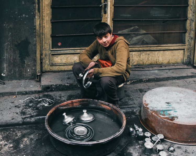 Boy Sitting And Cleaning Pots In Bucket