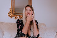 一位身穿花卉连衣裙的金发女子若有所思地坐在沙发上，背景是一面华丽的镜子