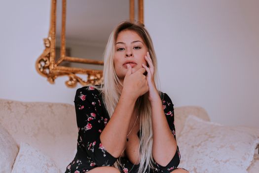 一位身穿花卉连衣裙的金发女子若有所思地坐在沙发上，背景是一面华丽的镜子