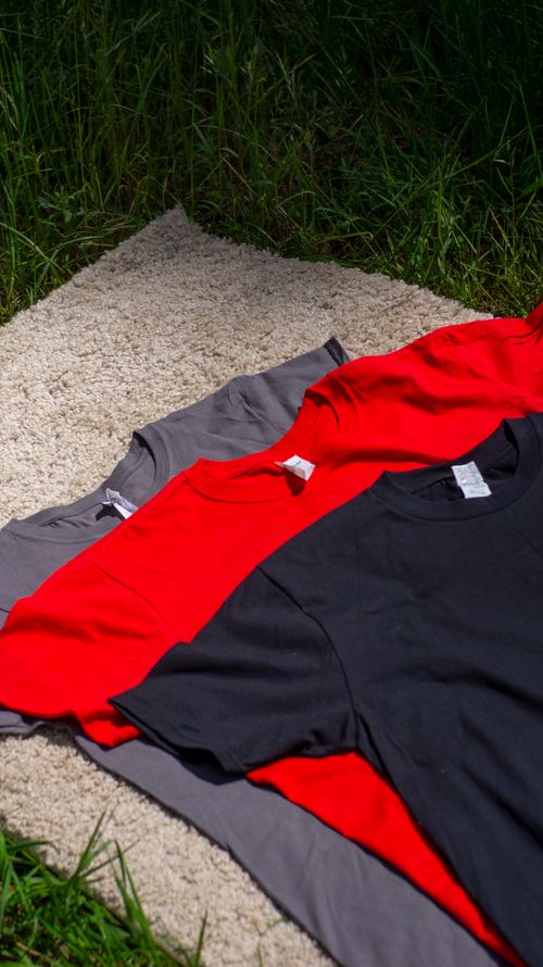 Free Three vibrant T-shirts in red, black, and grey laid out on a grass field in natural light. Stock Photo