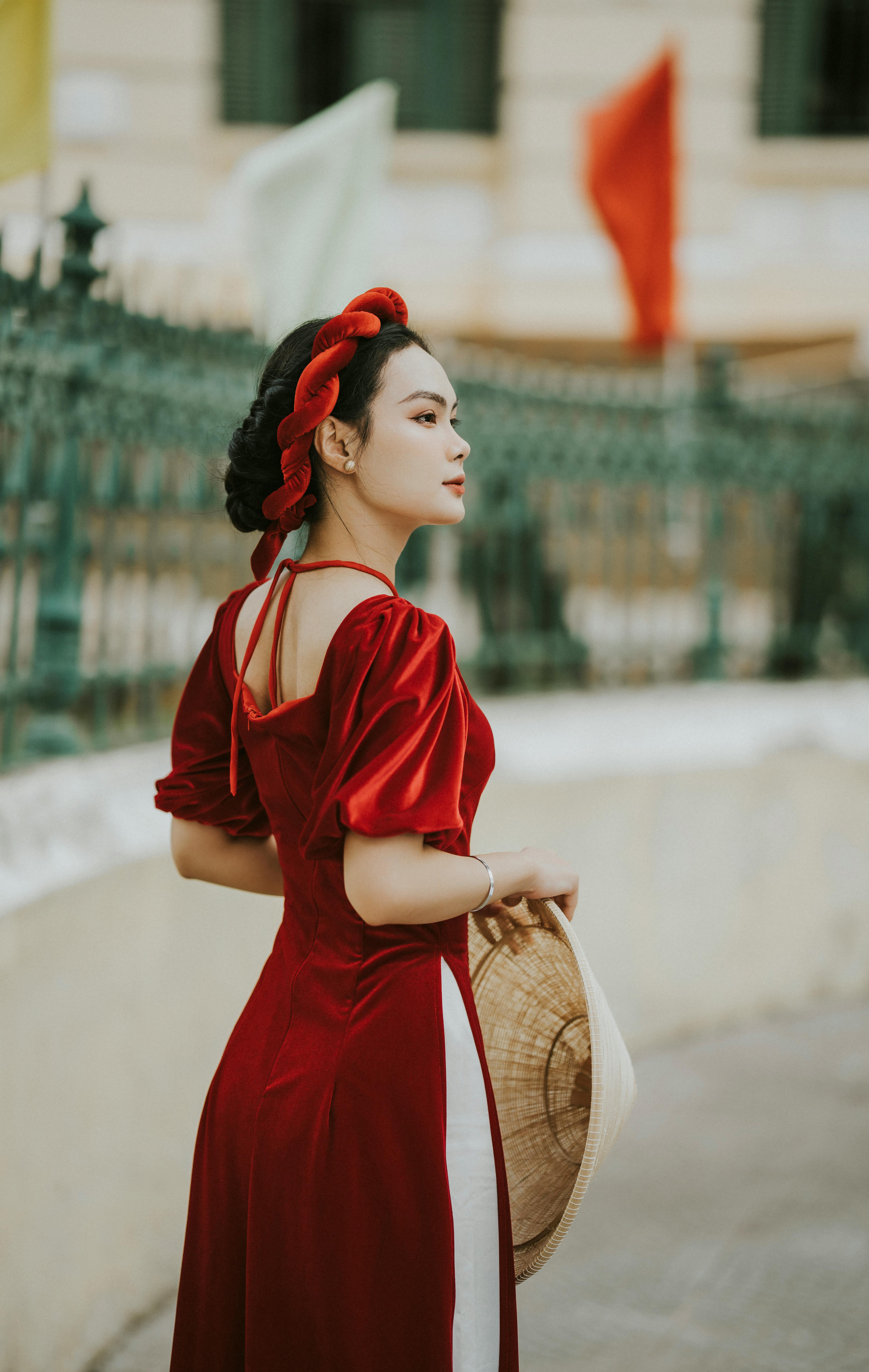 Asian woman in traditional attire holding conical hat, standing in urban setting.