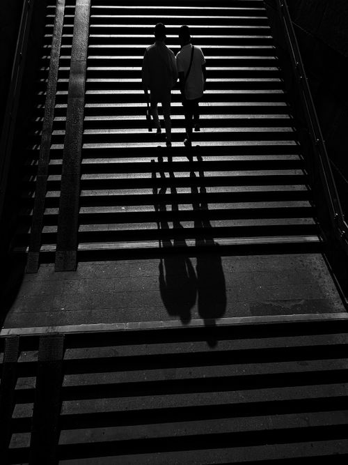 Free Black and white image of two silhouetted figures walking up an urban staircase with dramatic shadows. Stock Photo