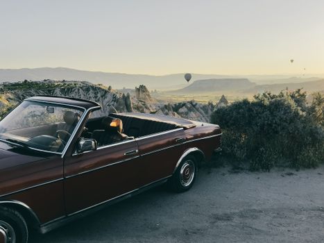 A classic brown convertible car parked in Göreme with hot air balloons rising at dawn.