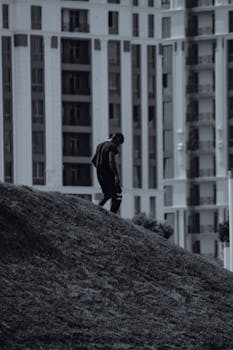 Black and white photo of a man walking up a grassy hill with urban buildings in the background.