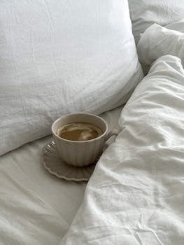 A cozy setup featuring a cup of coffee resting on white bed linens, evoking a sense of relaxation.