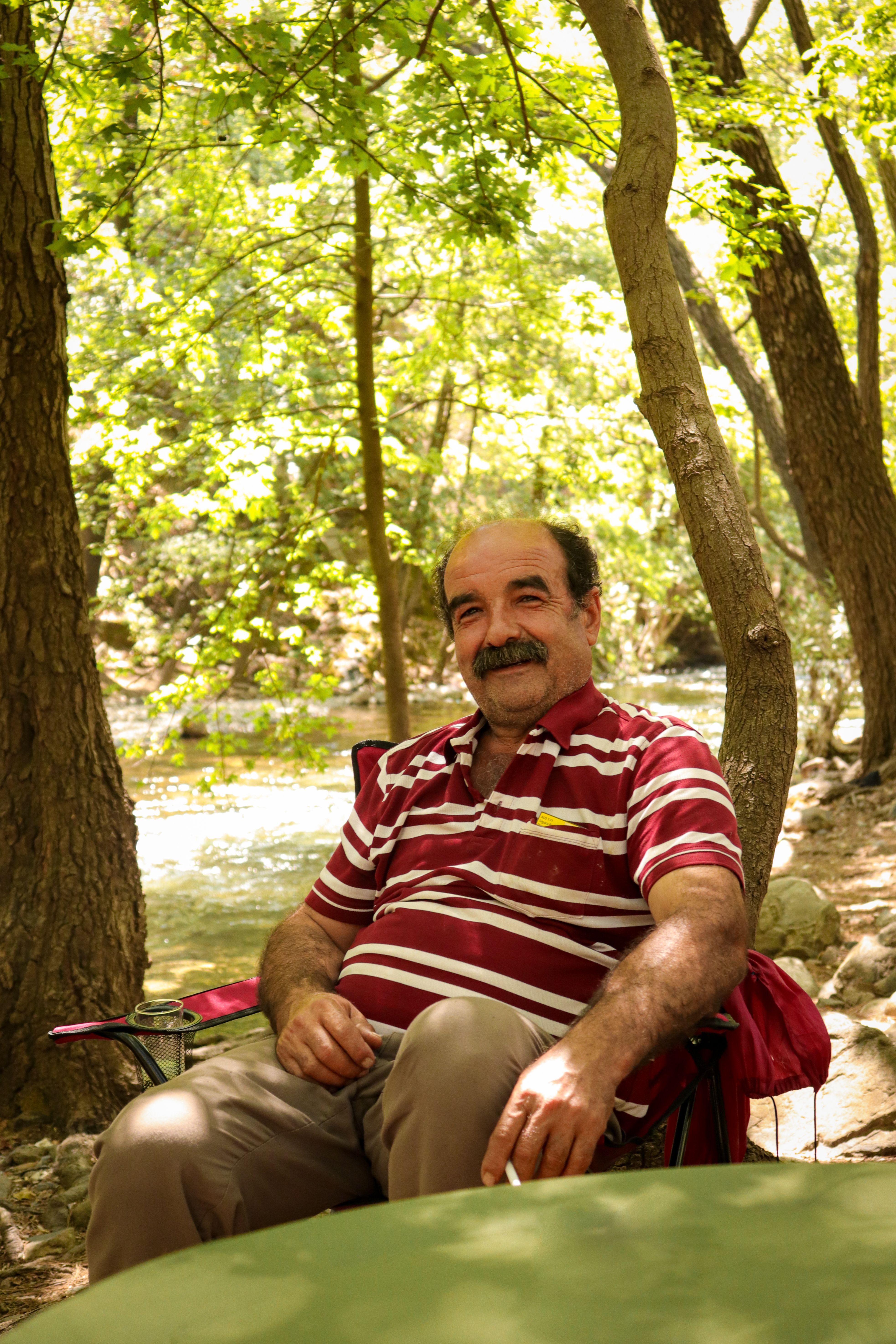 Elderly man with moustache sitting and smiling under trees in a tranquil Fethiye forest setting.