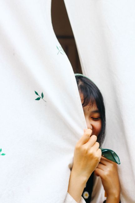 A woman playfully hides and smiles behind white curtains, creating a light-hearted moment indoors.