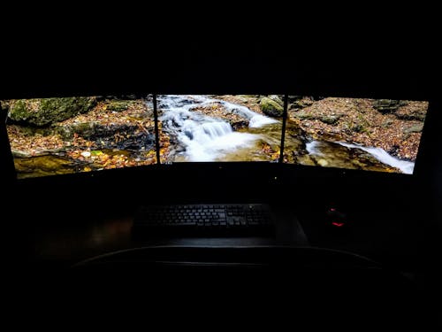 Modern computer with picturesque brook shown on screens placed on desk near keyboard and mouse in dark room