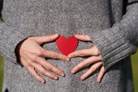 双手捧着一颗红色纸心，身穿灰色毛衣，象征着爱与关怀