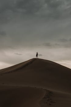 A solitary person stands atop a sand dune, silhouetted against an overcast sky.