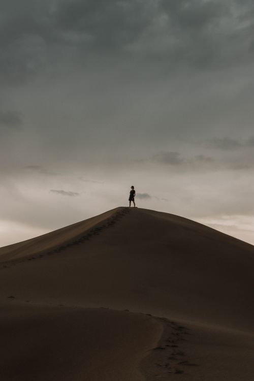 Free A solitary person stands atop a sand dune, silhouetted against an overcast sky. Stock Photo